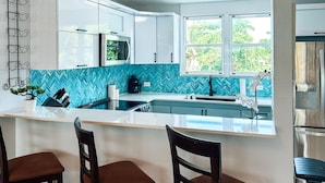 Una cocina moderna con armarios blancos, un salpicadero azul tipo espiga y un ventanal grande. Hay tres taburetes de bar de madera y un refrigerador de acero inoxidable.