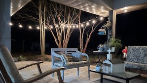 Eine Außenpatio-Terrasse mit einem Holzstuhl, einer gepolsterten Bank, einem Glastisch und einem gemusterten Sofa. Lichterketten und Blumentöpfe sind ebenfalls vorhanden.