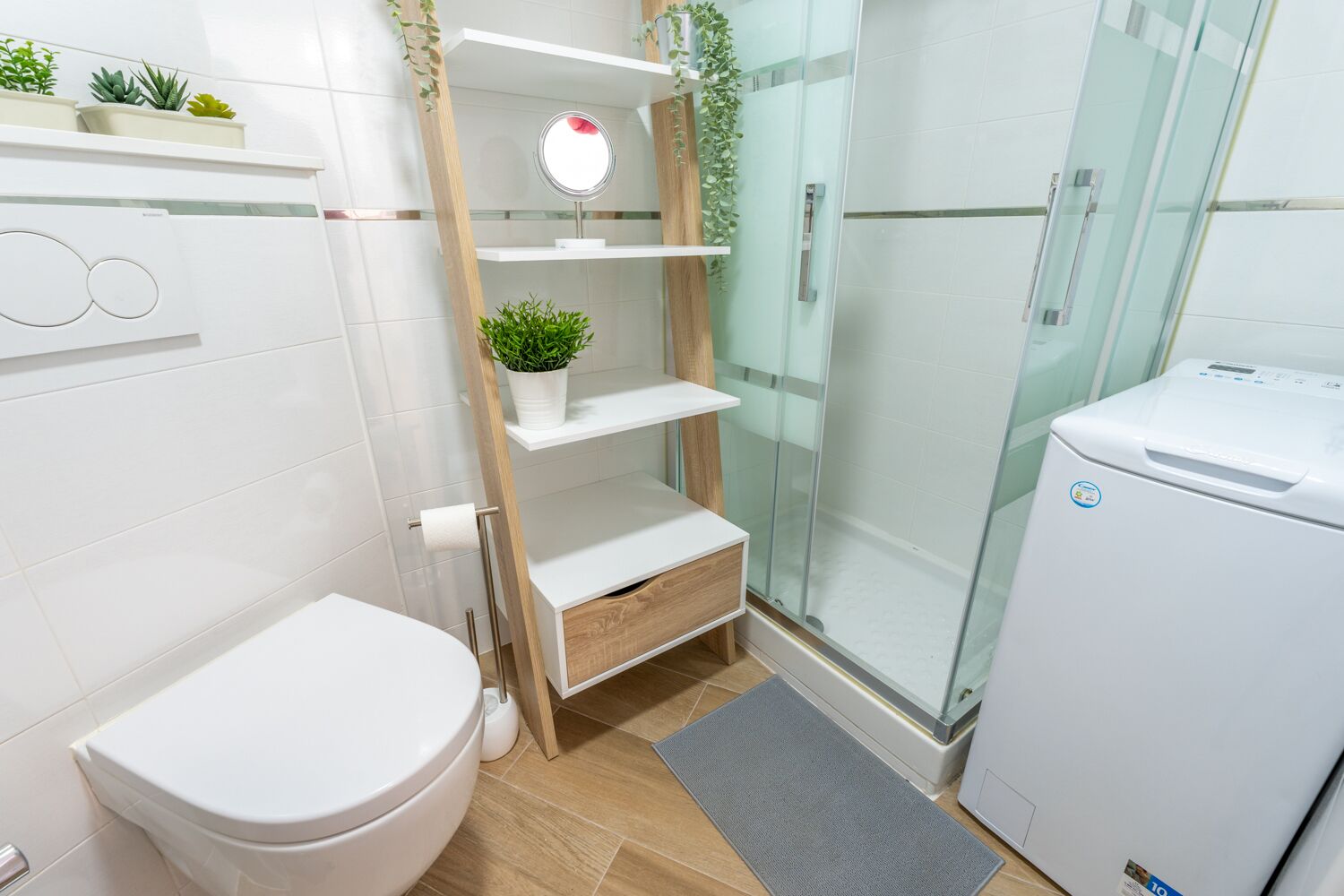A modern bathroom with a toilet, a glass shower enclosure, a built-in shelf with a mirror, and a small refrigerator.