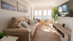 A modern living room with a beige sofa, a TV mounted on the wall, and a view of the sea through large windows.