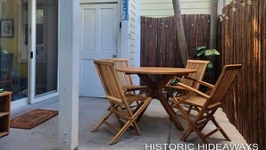 A wooden table and chairs set up in a covered outdoor area with a view of a white building in the background. - Cubana Casita (Key West)