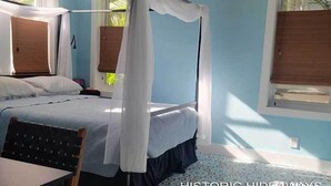 A bedroom with a four-poster bed, a wooden chair, and a window with a view of greenery. - Cubana Casita (Key West)