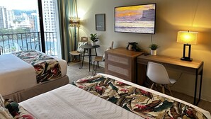 Habitación de hotel con dos camas, un escritorio, una silla, una mesita con una planta, una lámpara y un cuadro de un atardecer.
