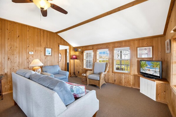 Una habitación acogedora con ventilador de techo, un sofá, un sillón de mimbre, televisión y paredes de madera.