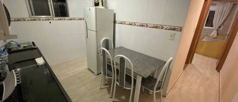 A compact kitchen with a refrigerator, a dining table with four chairs, a clock on the wall, and a window with a curtain.