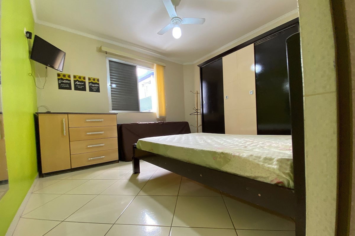 A bedroom with a bed, a wardrobe, a desk with a chair, and a television on the wall.