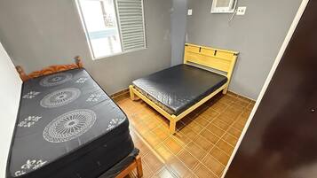 A small room with two beds, a window with blinds, an air conditioning unit, and a tiled floor.