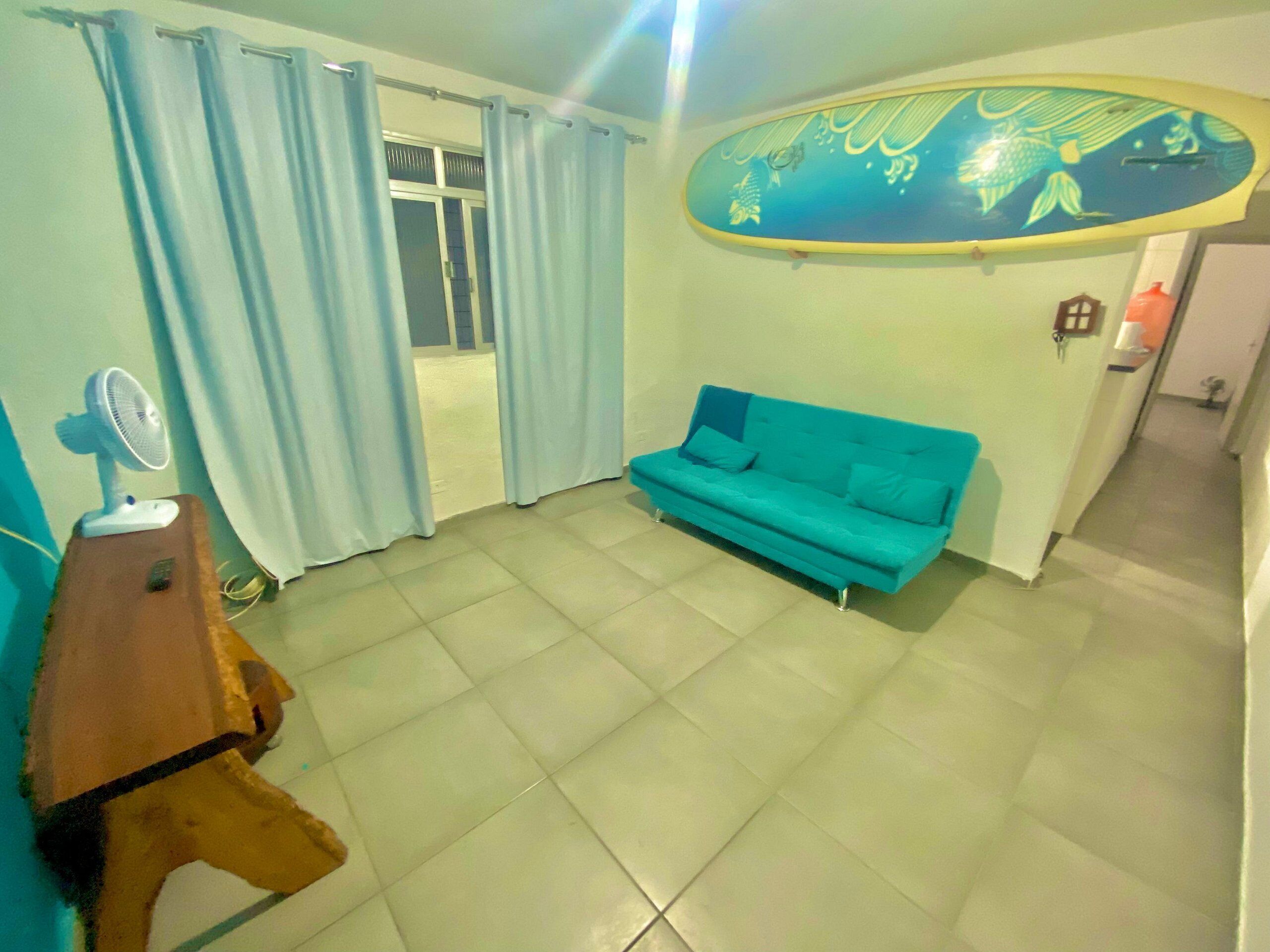 Un salon avec un canapé de couleur turquoise, une petite table sur laquelle se trouve un ventilateur, et une décoration murale représentant une planche de surf.