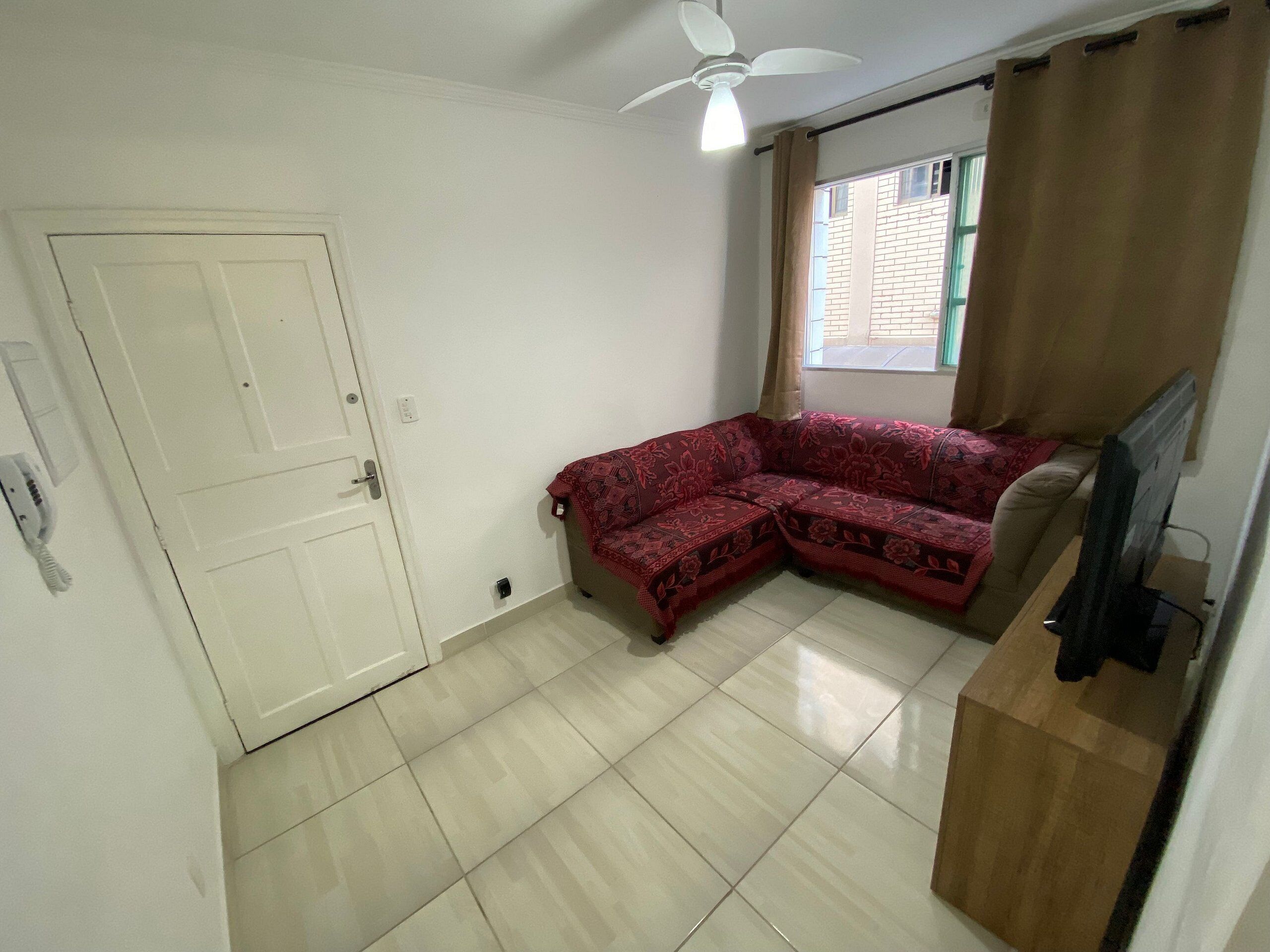 Une pièce avec une porte blanche, un ventilateur de plafond, une fenêtre avec des rideaux et un canapé rouge à motifs.