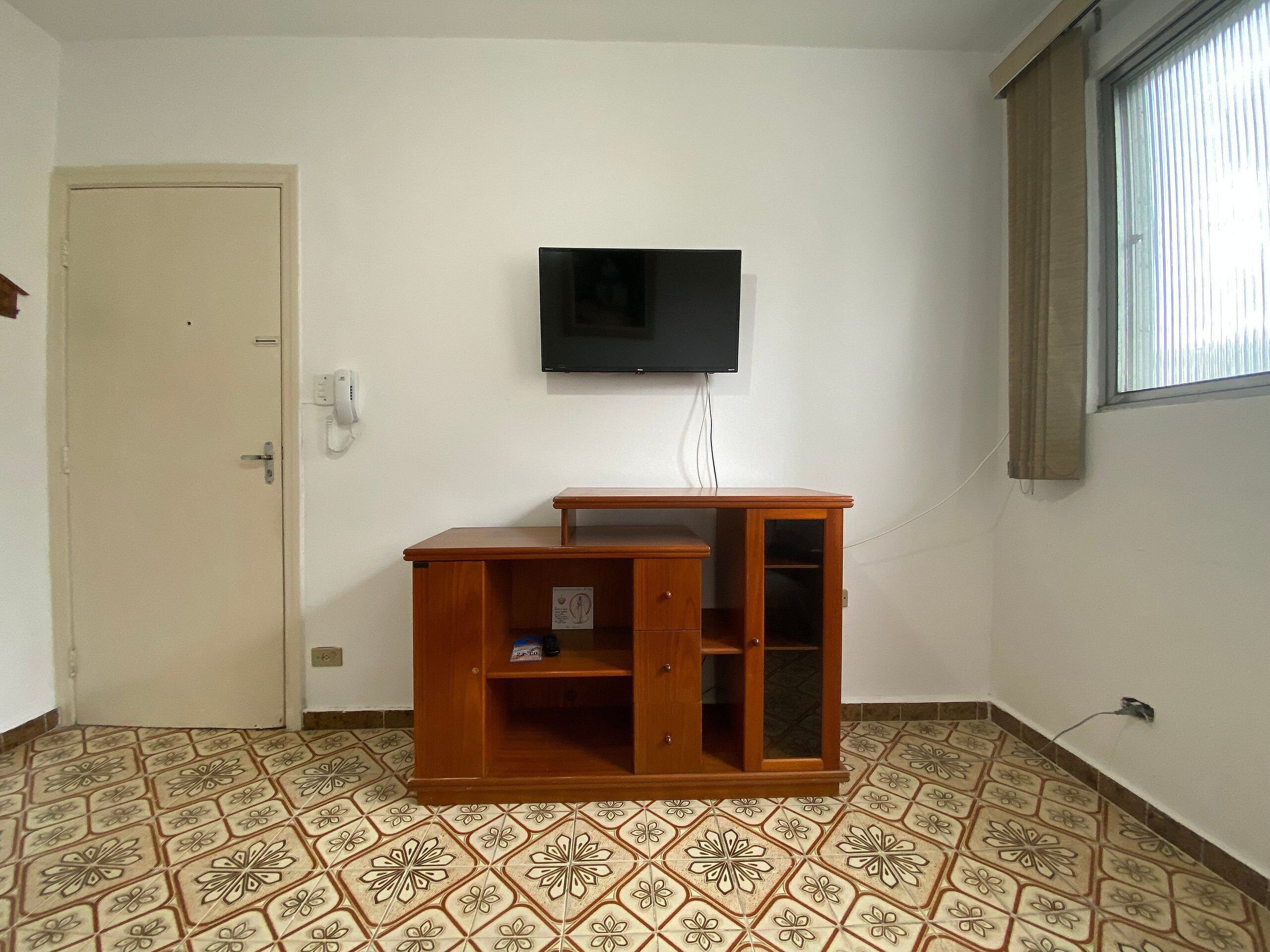 A room with a wooden desk, a mounted television, patterned floor tiles, a door, and a window with blinds.