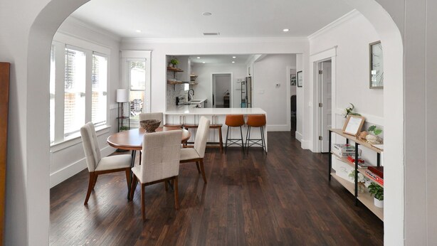 A spacious kitchen with a central island, wooden flooring, and a white archway leading to a dining area with a round table and chairs.