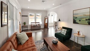 A spacious living room with a brown leather sofa, a green armchair, and a wooden coffee table. There is a painting on the wall and a small round table with candles.