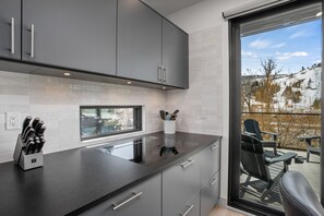 Une cuisine moderne avec des meubles gris, un plan de travail noir et une fenêtre donnant sur un espace extérieur enneigé, agrémenté de fauteuils Adirondack.