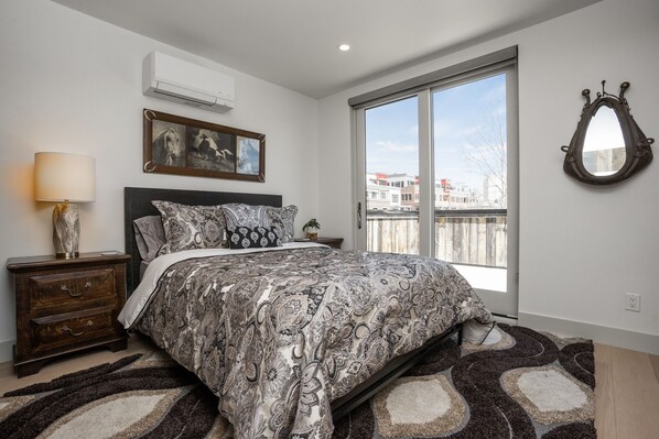 Une chambre à coucher avec un grand lit, une table de chevet, une lampe, un tableau encadré et une vue sur les immebles par la fenêtre.