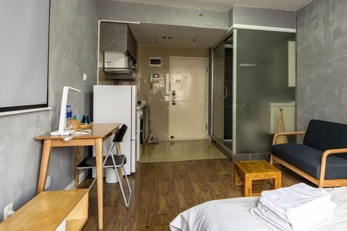 A compact hotel room with a bed, a desk, a chair, a small table, a sofa, and a glass door leading to a bathroom.