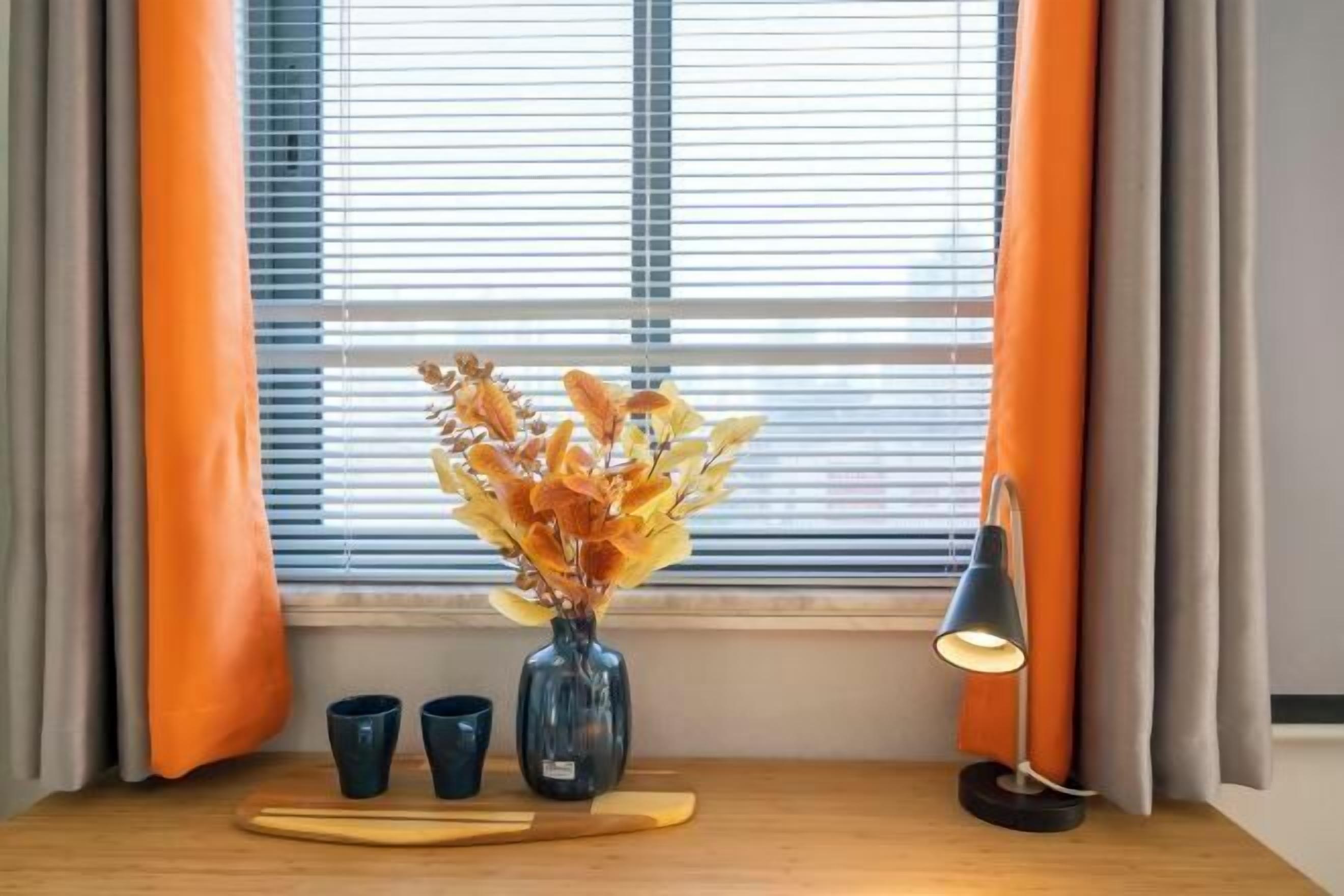 Ein Zimmer mit Fenster, orangefarbenen Vorhängen, einer Vase mit getrockneten Blumen, zwei Tassen, einem Holztablett und einer Schreibtischlampe.