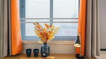 Ein Zimmer mit Fenster, orangefarbenen Vorhängen, einer Vase mit getrockneten Blumen, zwei Tassen, einem Holztablett und einer Schreibtischlampe.