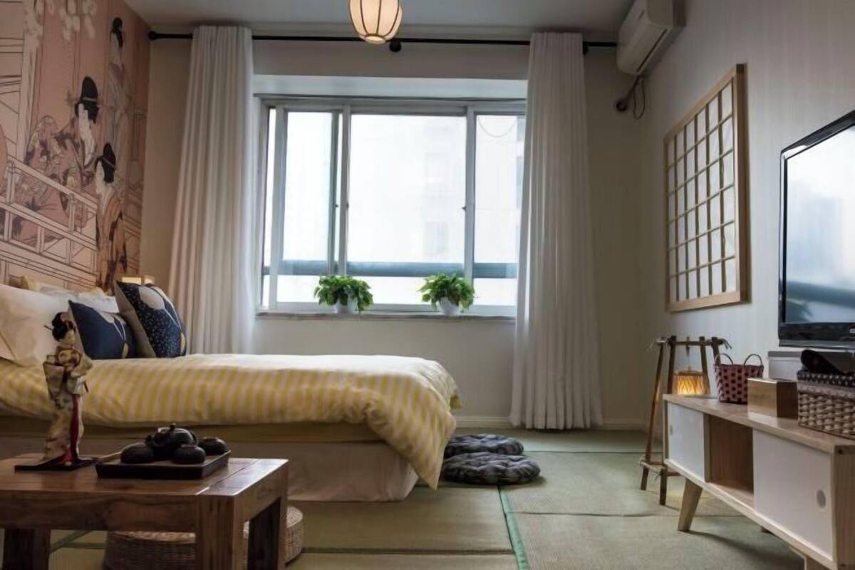 A room with a bed, a window with plants, a TV, and a traditional Japanese-style desk.