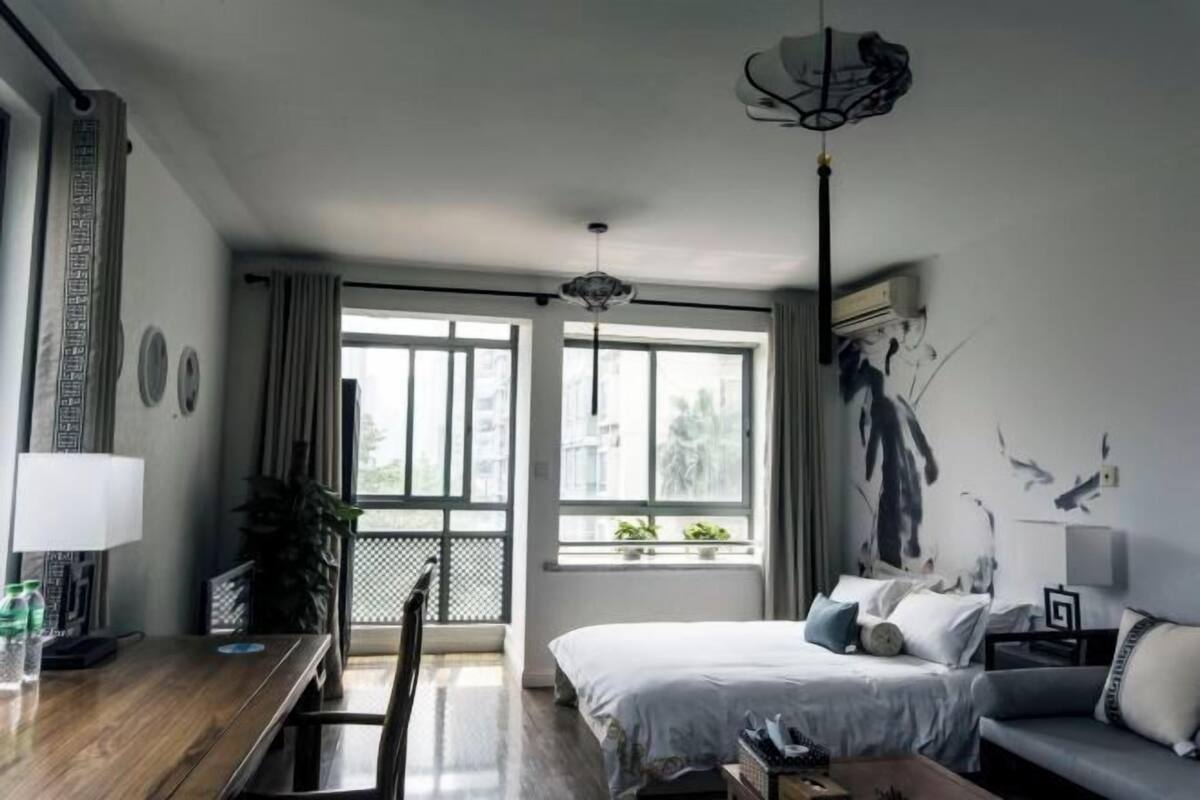 A modern hotel room with a large bed, a desk, and a sofa. There are windows with curtains, a ceiling fan, and a wall with a bird mural.