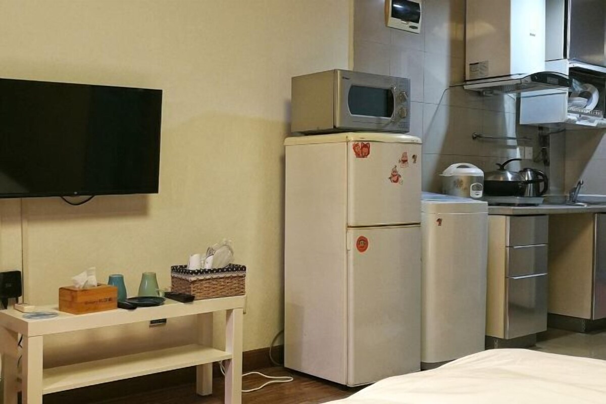 A compact hotel room with a kitchenette, a flat-screen TV, a small table with toiletries, and a bed.