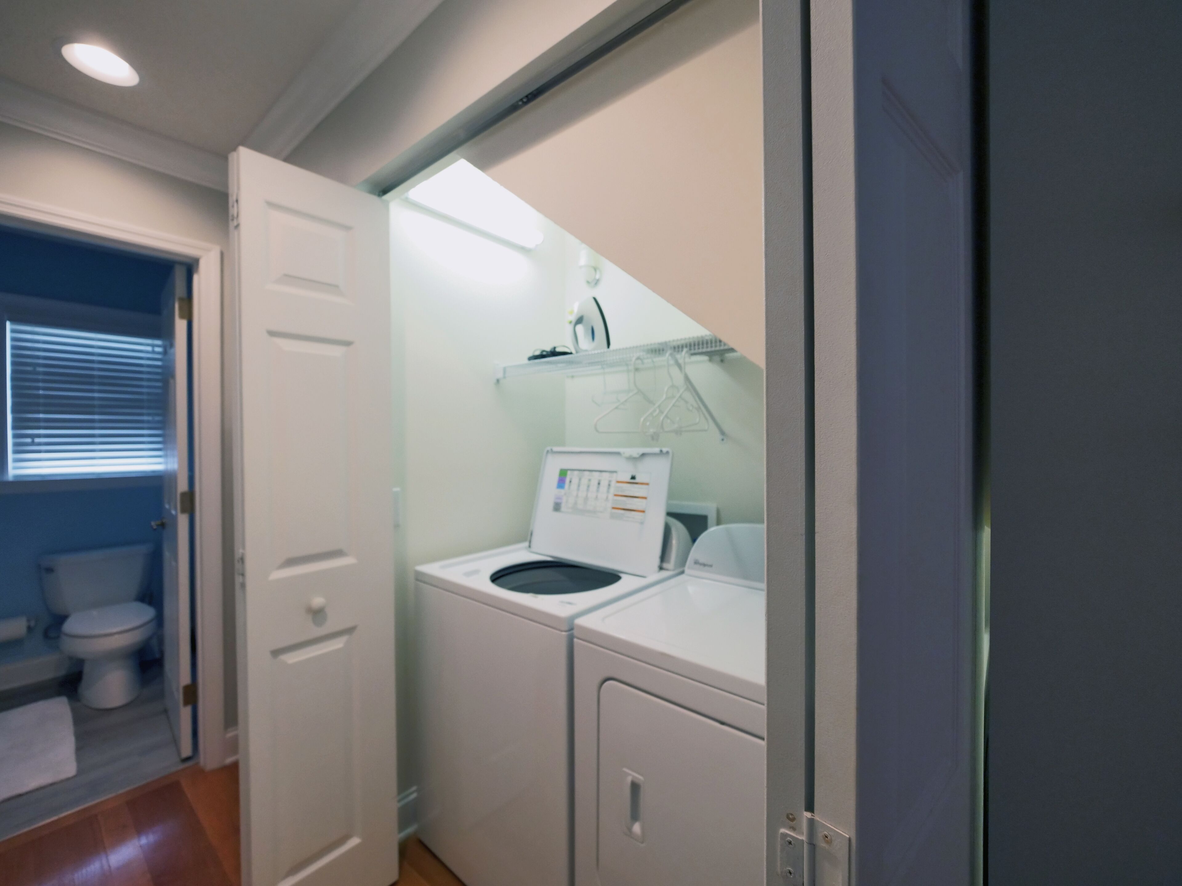 Een wasruimte met een wasmachine en droger, een klein raam met jaloezieën, en een deur naar een andere kamer.