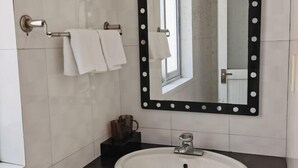 Un baño con lavamanos blanco, un espejo con marco negro y dos toallas colgadas en un perchero.