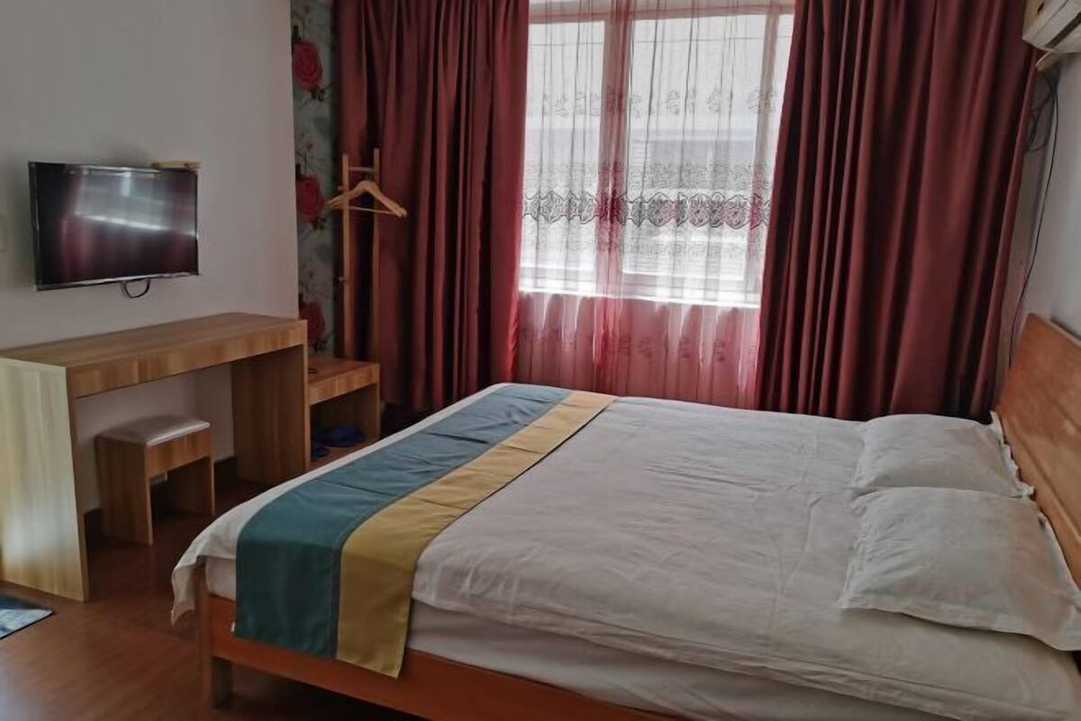 Une chambre à coucher avec un lit, un téléviseur fixé au mur, un bureau avec une chaise et une fenêtre avec des rideaux.
