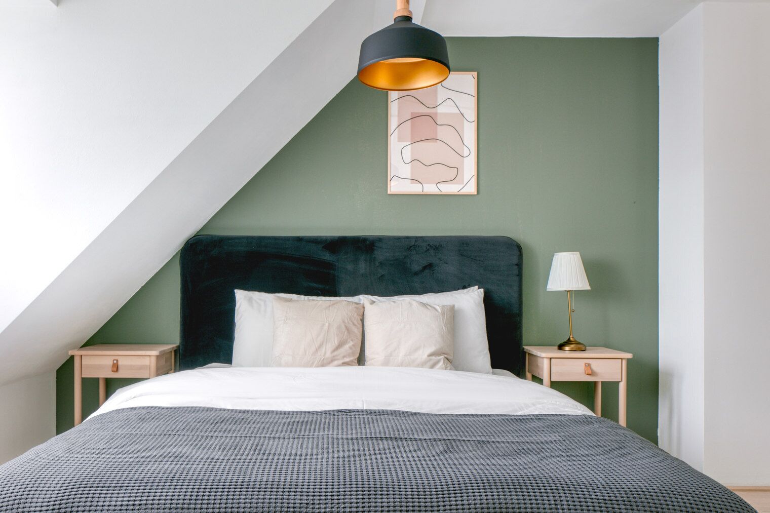 Een slaapkamer met een donkergroen hoofdbord, een witte spreidek en een deken met een hondentand-patroon. Er hangt een abstract kunstwerk aan de muur en een vloerlamp staat op een nachtkastje.