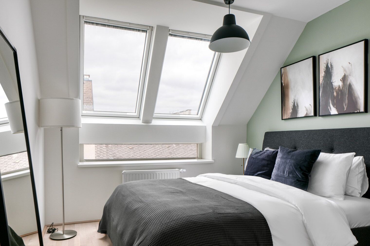 Un dormitorio moderno con tragaluz, una cama con ropa de cama blanca y oscura, una lámpara de pie y una obra de arte en la pared.