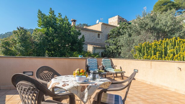 Une terrasse avec une table dressée pour deux personnes, entourée de chaises, et une vue sur un bâtiment en pierre et des arbres en arrière-plan.
