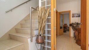 Un escalier avec une rampe métallique, un pot en cuivre contenant des plantes séchées, posé sur un support, une porte en bois et un miroir qui reflète un couloir orné d’un tableau.
