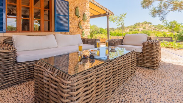 Uma área de estar externa com móveis de vime e uma mesa de vidro, em frente a um edifício de pedra com vista para o campo.