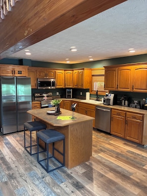 A kitchen with wooden cabinets, stainless steel appliances, a central island with stools, and a coffee station.