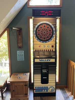 Eine Dartscheibe mit digitaler Punktetafel darüber, eine gerahmte Uhr und ein Schild mit den Aufschriften 'Lake' und 'Valley Darts' an einer Wand.