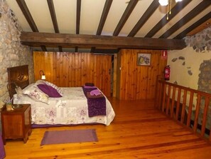 A bedroom with a wooden floor, a bed with purple bedding, a nightstand, a wooden headboard, and a stone wall.