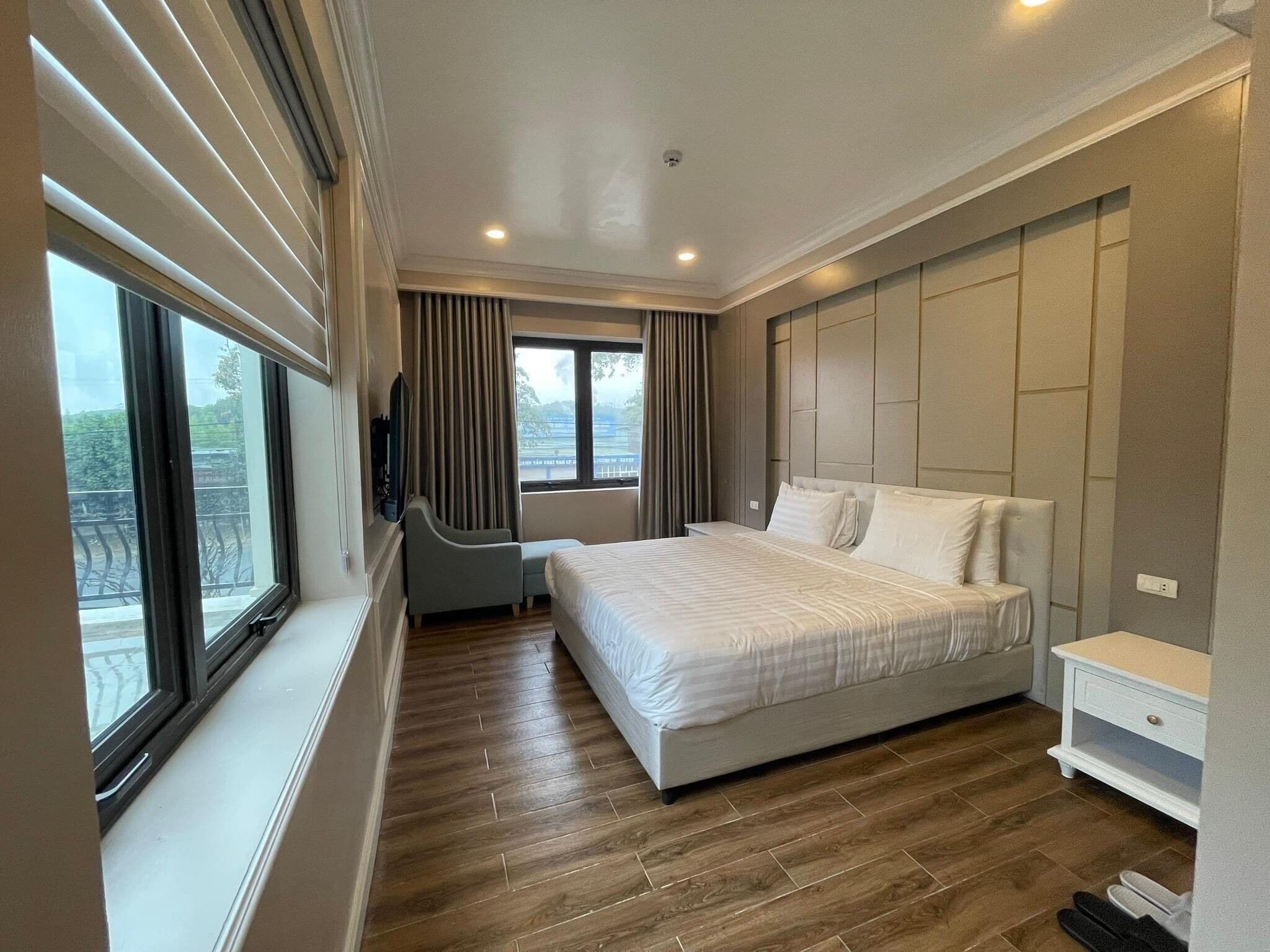 A modern hotel room with a large bed, a desk, and a chair. There are windows with curtains, a view of the outdoors, and a ceiling light.