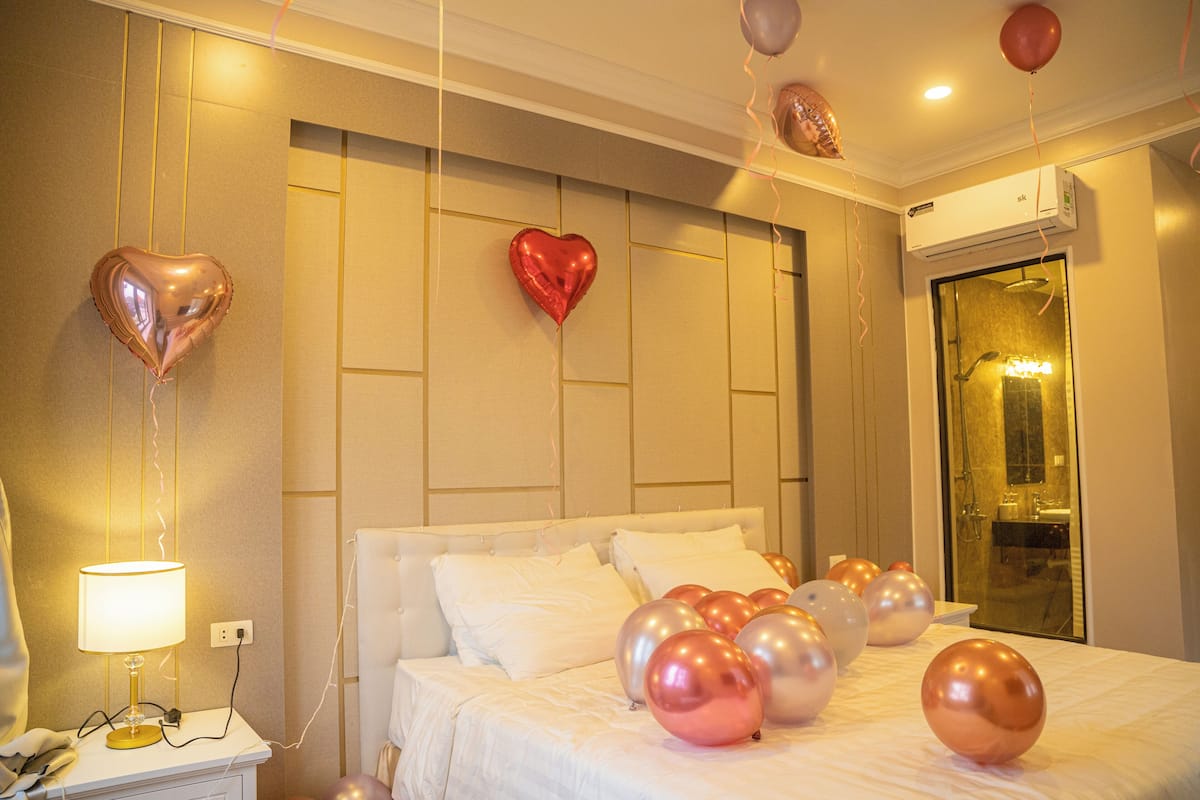 A hotel room with a bed, a bedside table with a lamp, and balloons on the bed.