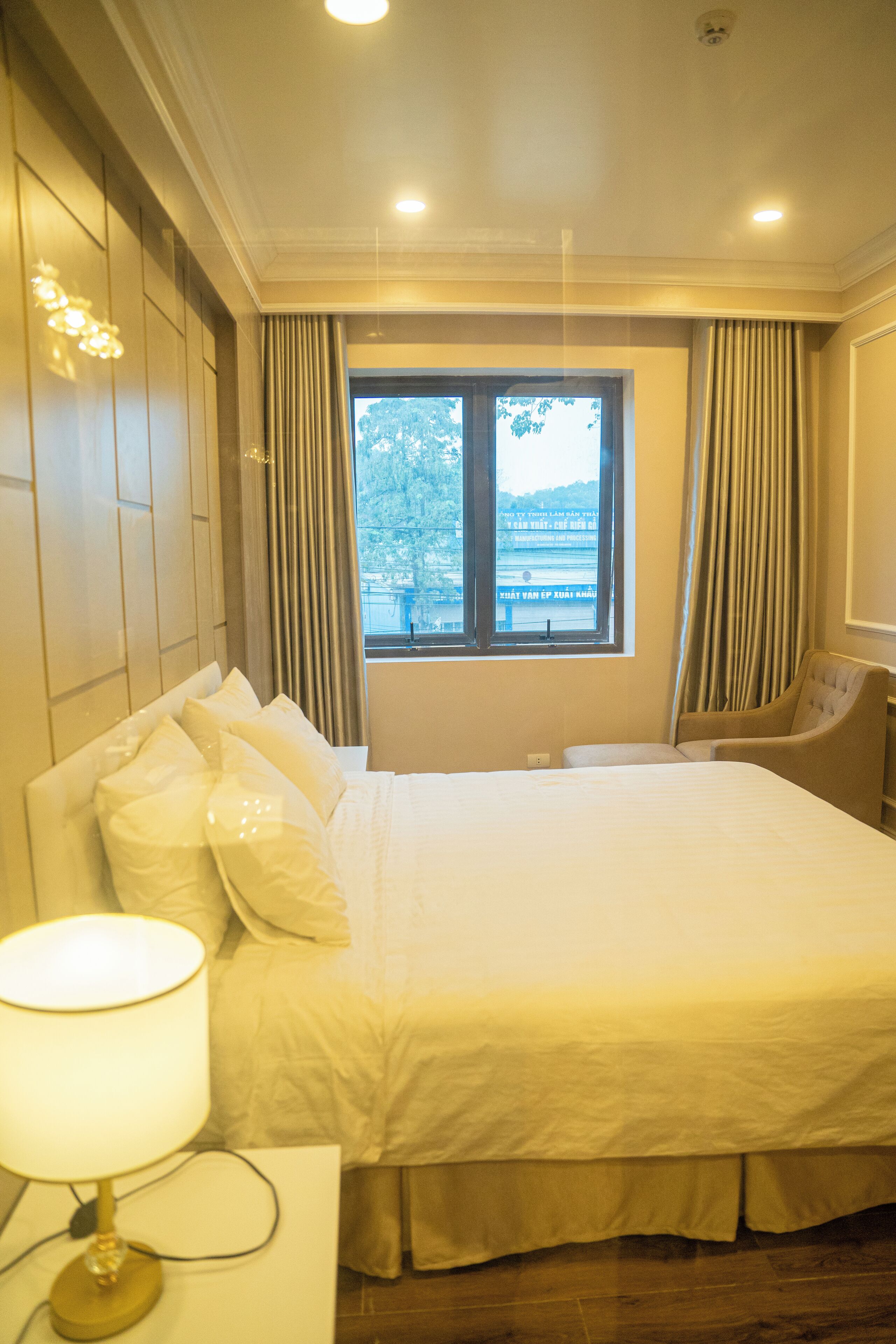 A hotel room with a large bed, a nightstand with a lamp, a chair, and a window with curtains.