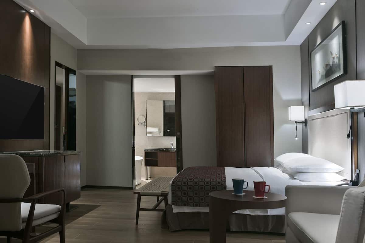 A hotel room with a bed, a desk, a chair, a TV, a bedside table with two cups, and a wardrobe.