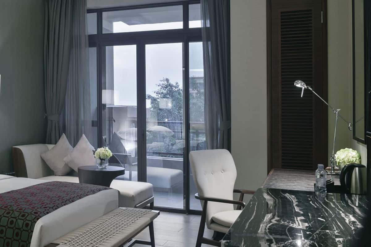 A hotel room with a bed, a desk, a chair, a table, a lamp, a window with curtains, and a view of the outdoors.
