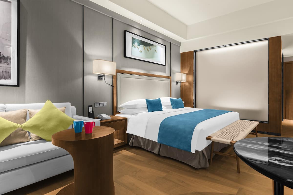 A modern hotel room with a bed, bedside table, lamp, and a view of the city.