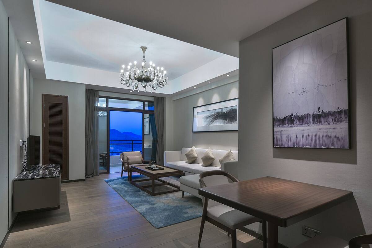 A modern living room with a sofa, coffee table, and a chandelier. There is a large window with a view of the city and mountains.