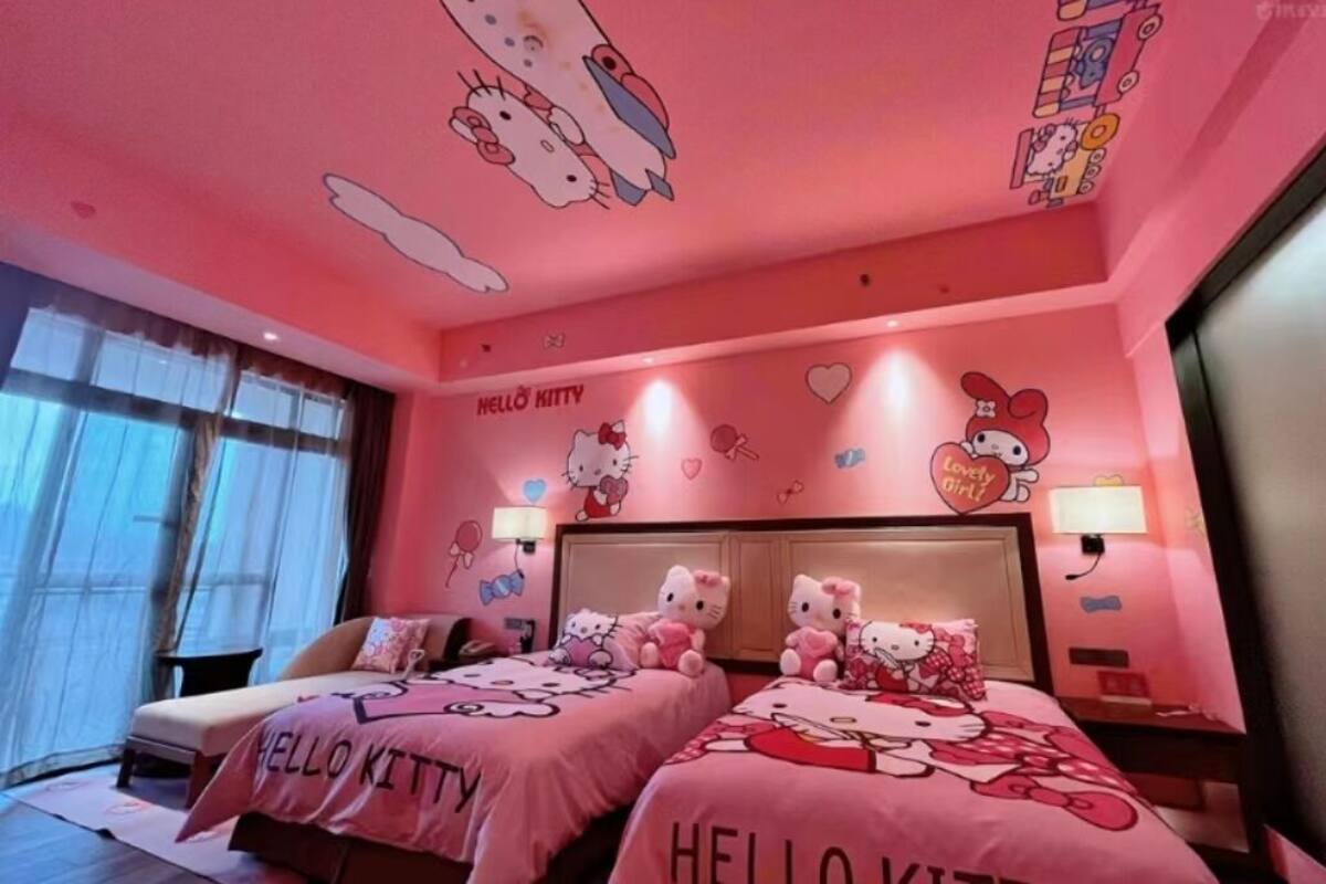 A pink-themed room with two beds, each adorned with Hello Kitty bedding and pillows. The walls feature Hello Kitty decorations, and there are two bedside lamps.