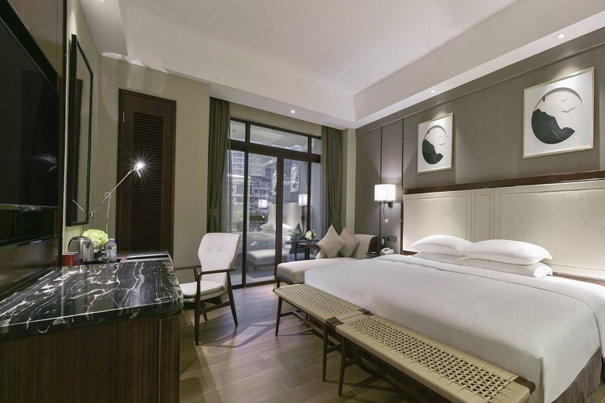 A hotel room with a large bed, a desk with a chair, a bench, a TV, and a window with a view of the city.