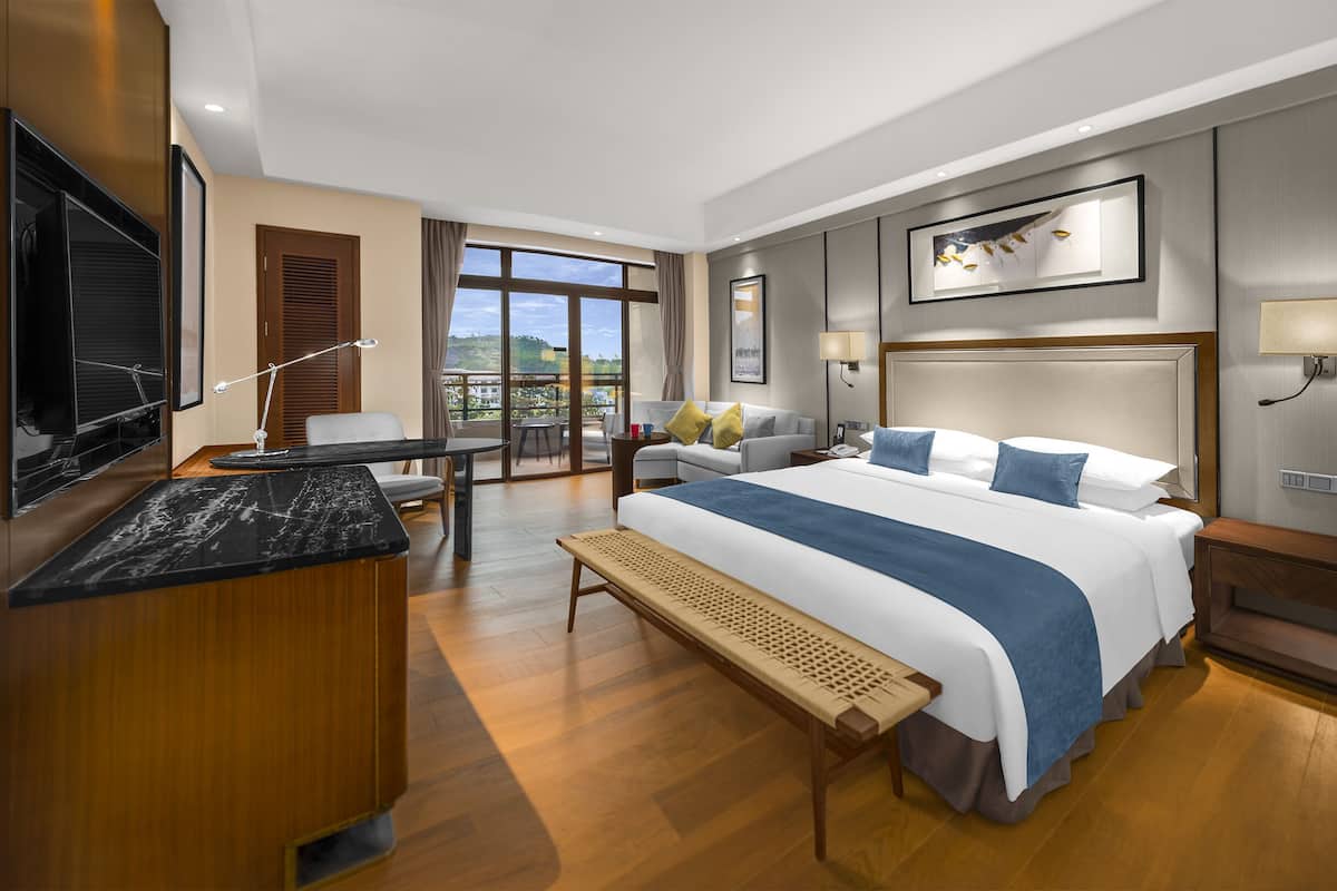 A hotel room with a large bed, a desk, a TV, and a balcony with a view.
