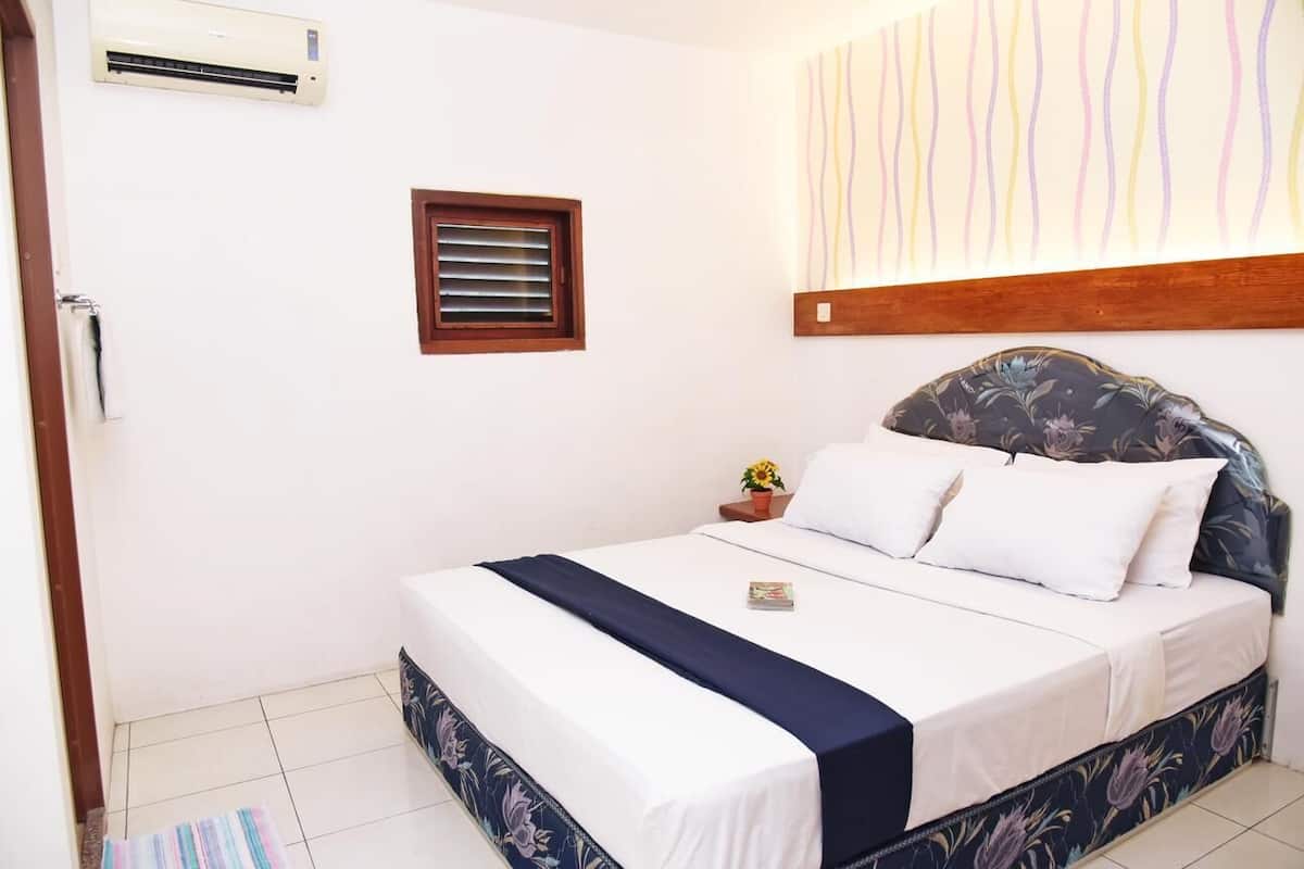 Een slaapkamer met een bed, een nachtkastje met een plant, een raam met jaloezieën en een airconditioning aan de muur.