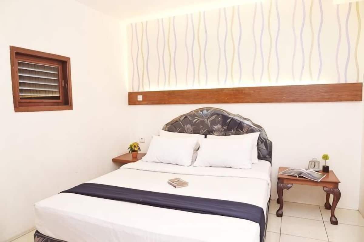 Een slaapkamer met een bed, een nachtkastje, een wandplank en een raam met jaloezieën.