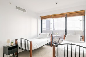 Two single beds with wooden frames and white bedding, a small dining table and chair, and a bedside table with a lamp.