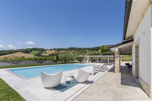 A swimming pool with white chairs and a covered area, surrounded by a hilly landscape. - Villa Ada by MMega Villas  (Lapedona)