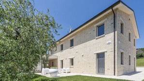 A two-story building with a flat roof, a brick facade, and a covered outdoor seating area with white chairs and a large planter. - Villa Ada by MMega Villas  (Lapedona)